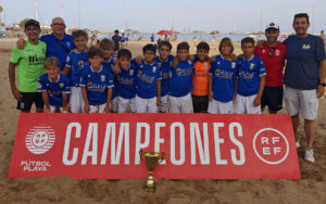 Youth soccer team on the beach posing with a large red 'CAMPEONES' banner and a trophy in front.