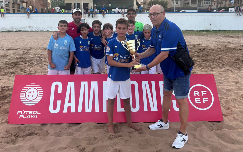 Group of young soccer players in blue uniforms posing on a sandy field as a coach hands a trophy to the front boy.