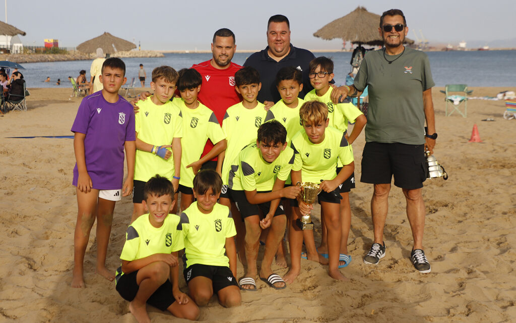 Youth soccer team on a sandy beach posing with a trophy, flanked by two coaches, wearing neon jerseys and shorts by the sea.