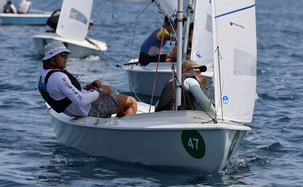 Small sailboat race: two sailors in a dinghy numbered 47, steering through blue water with other boats in the background; one crew member wears a white hat and sunglasses.