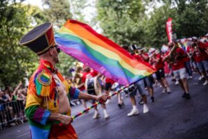 Performer in a colorful rainbow costume marching in a Pride parade while holding a rainbow flag on a striped staff.
