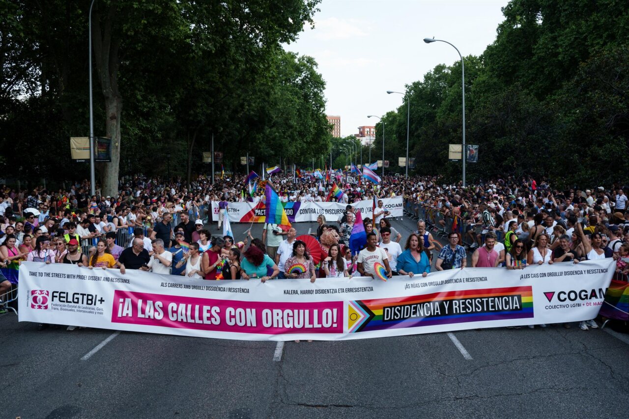 A large LGBTQ+ pride march on a city street with rainbow flags and banners reading slogans like '¡A las calles con orgullo!' and 'Disidencia y Resistencia.'