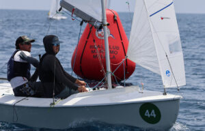 Two sailors in a white dinghy with a green 44 badge on the bow navigate near a large red buoy labeled for a regatta, out on calm blue water.