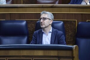 Man in a navy blazer and light shirt sits in a formal council chamber, gazing to his left with a thoughtful expression.
