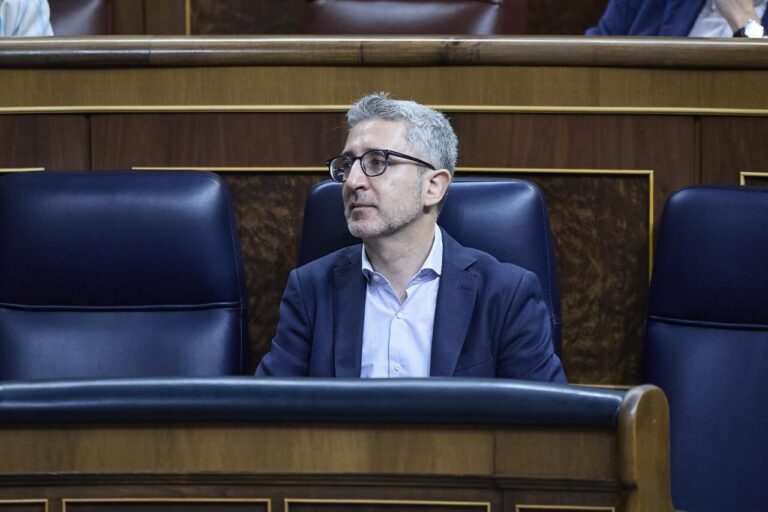Man in a navy blazer and light shirt sits in a formal council chamber, gazing to his left with a thoughtful expression.