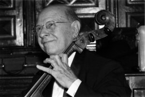 Older man in a suit playing a cello indoors, smiling slightly.