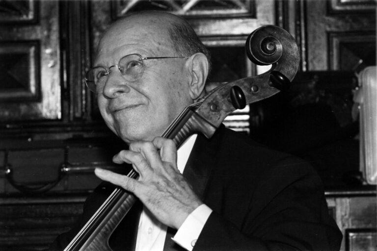 Older man in a suit playing a cello indoors, smiling slightly.