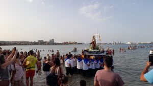 Crowd gathered at a beach watching a religious float with a statue of a saint being carried through shallow water near shore.