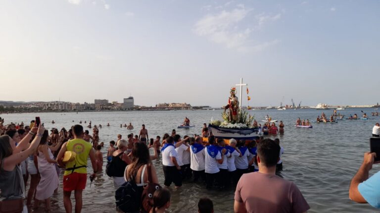 Crowd gathered at a beach watching a religious float with a statue of a saint being carried through shallow water near shore.