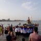Crowd gathered at a beach watching a religious float with a statue of a saint being carried through shallow water near shore.