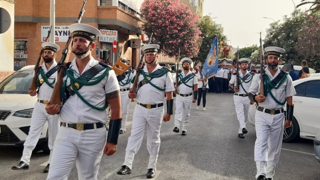 Sailors in white uniforms march in a street parade, rifles shouldered, green cords draped over their chests, with spectators and a blue flag in the background.