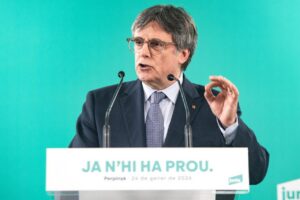 Man in a suit and glasses speaks at a podium with a teal background, gesturing with his right hand.