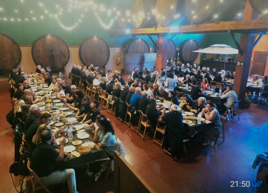 Large group of people seated at long communal tables for a dinner in a winery-style hall with large wooden barrels and string lights above.
