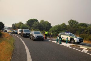 Line of cars stuck in a traffic jam on a rural highway as two officers in high-visibility vests direct traffic near a Guardia Civil vehicle on the right.