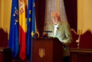 Older man in a light green blazer delivering a speech at a wooden podium, with European and national flags behind him in a formal room.