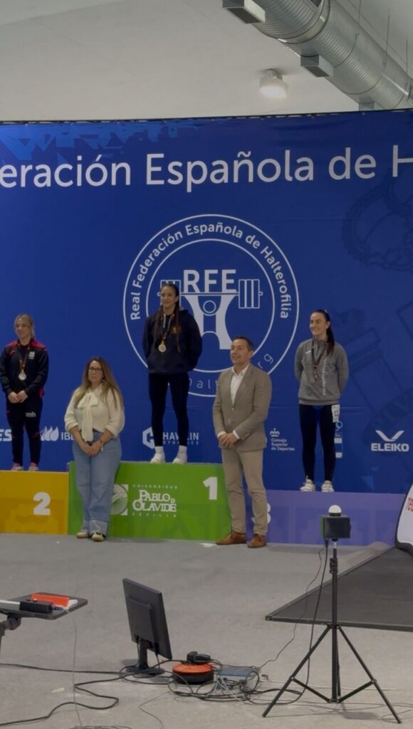 Award ceremony on a podium with 1st, 2nd, and 3rd place platforms; a man in a suit stands nearby as athletes wear medals against a blue federation banner.