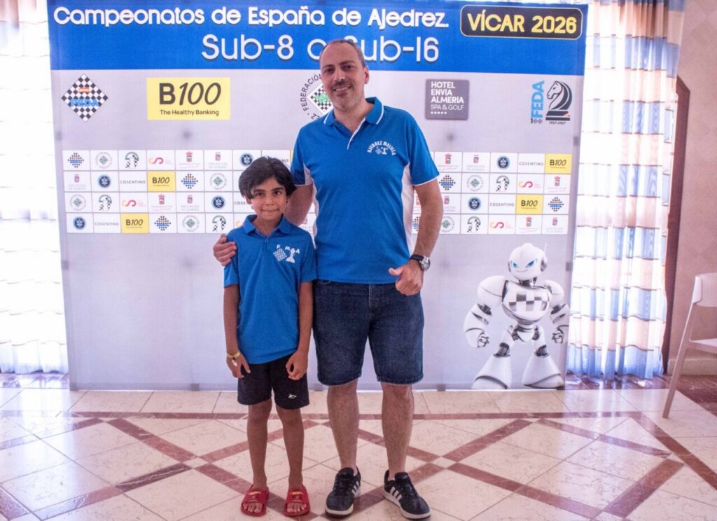 Man and boy in matching blue shirts pose for a photo in front of a chess championship banner (Spain, Sub-8/Sub-16).