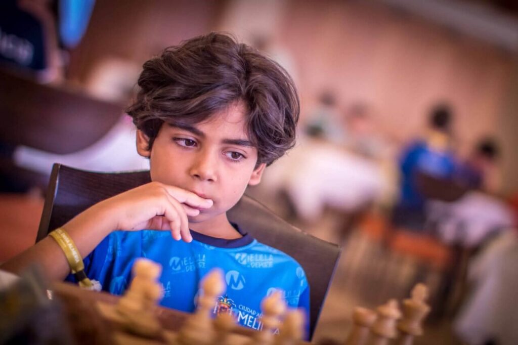 Boy with brown hair in a blue shirt sits at a chessboard, deep in thought as he considers his next move.