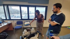 Woman demonstrates a tablet on a stand to a baby in a stroller while a man watches in a bright office. In the background, a window shows a parking lot.