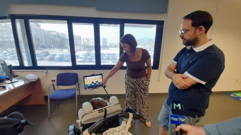 Woman demonstrates a tablet on a stand to a baby in a stroller while a man watches in a bright office. In the background, a window shows a parking lot.