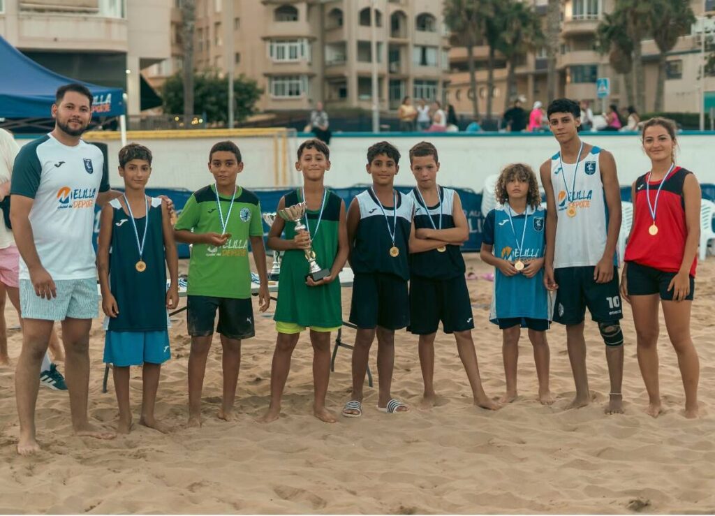 Group of young athletes in team jerseys stand on a sandy beach, wearing medals; one child holds a trophy while a coach stands on the far left after a competition.