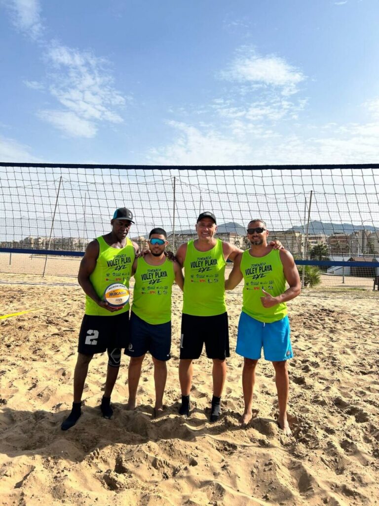 Four men in neon green tank tops stand arm in arm on a sandy beach volleyball court, with a net behind them and a clear blue sky; the man on the far left is holding a volleyball.
