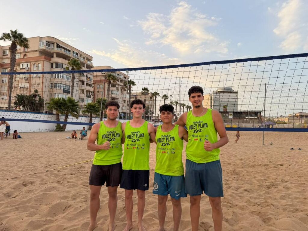 Four young men in neon green volleyball jerseys pose together on a sandy beach volleyball court, with palm trees and buildings in the background.