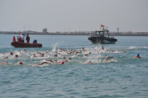 Group of swimmers doing a sea race near a harbor, with a red inflatable rescue boat on the left and a black support boat on the right.