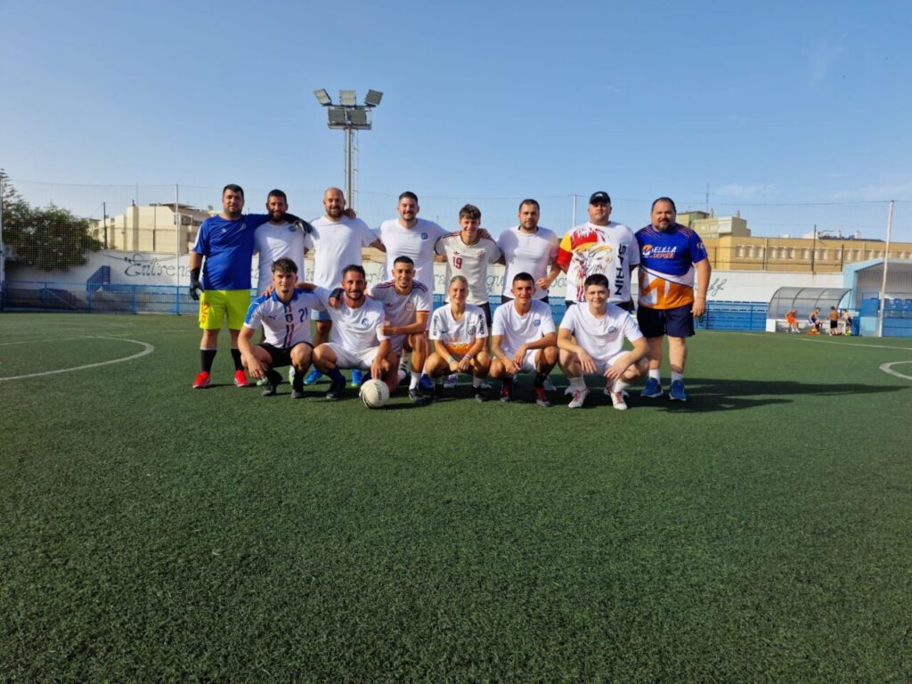 Group of soccer players posing for a team photo on an outdoor field, white kits with one blue-jersey goalkeeper, stadium in background.