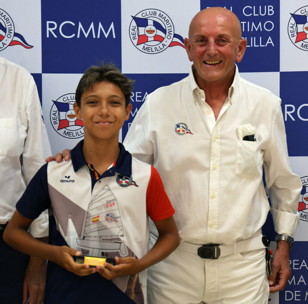 Boy smiles while holding a trophy, standing with an older man in a white shirt at a club awards backdrop (RCMM logo).