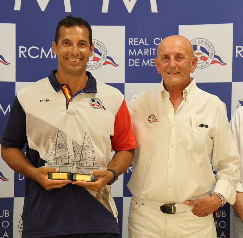 Two men pose at a sailing club awards event; the man on the left holds two model sailboats trophies and smiles at the camera.