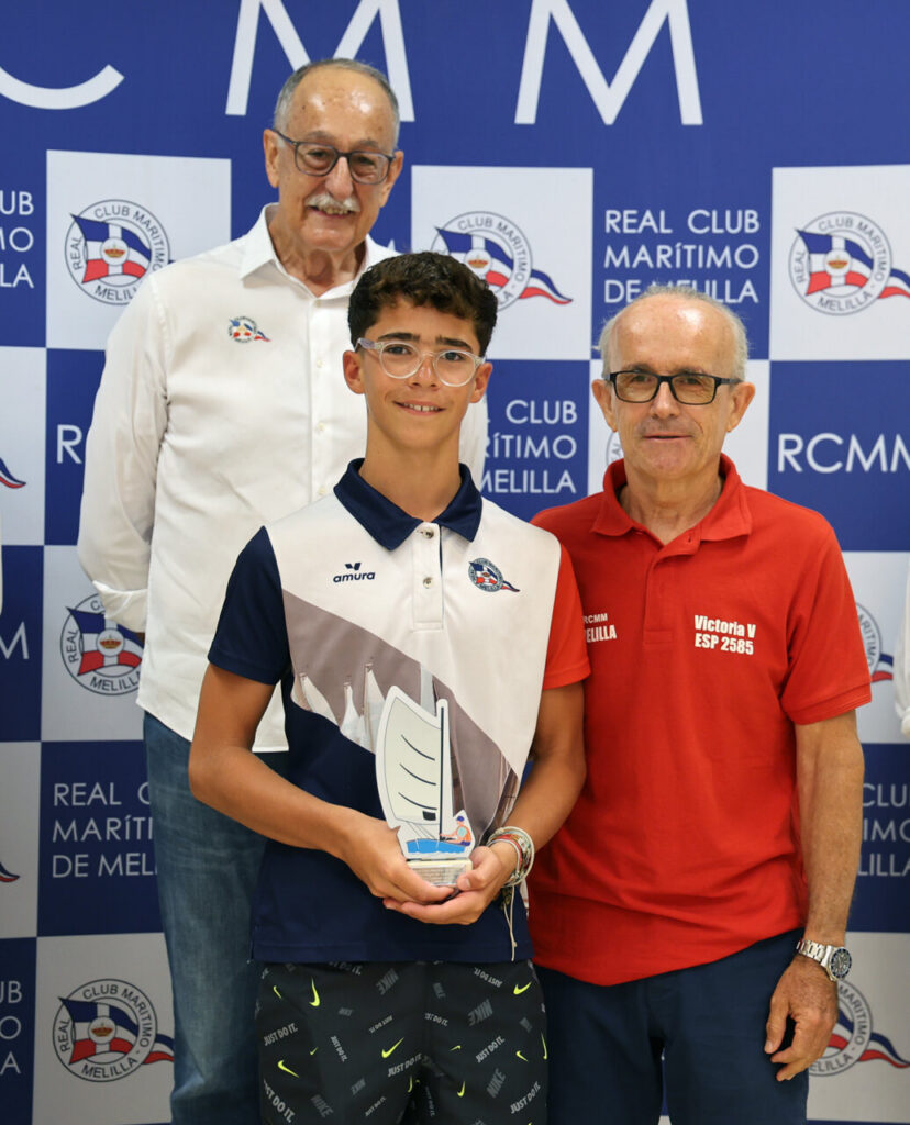 Young boy with glasses holding a sailboat trophy, flanked by two men at a Real Club Marítimo Melilla event backdrop.