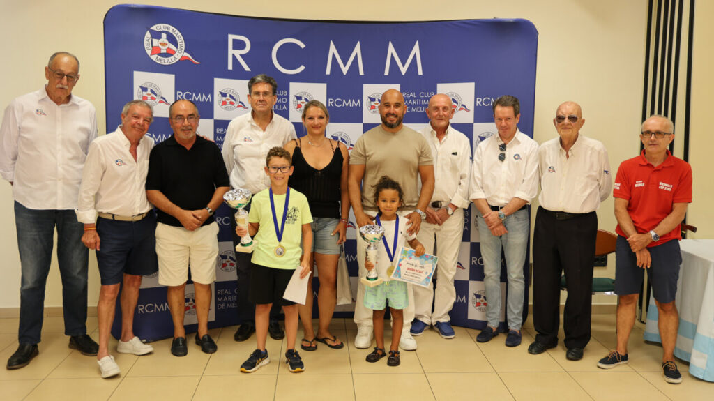 Group of adults and two children posing with trophies at an RCMM event backdrop featuring logos and branding.