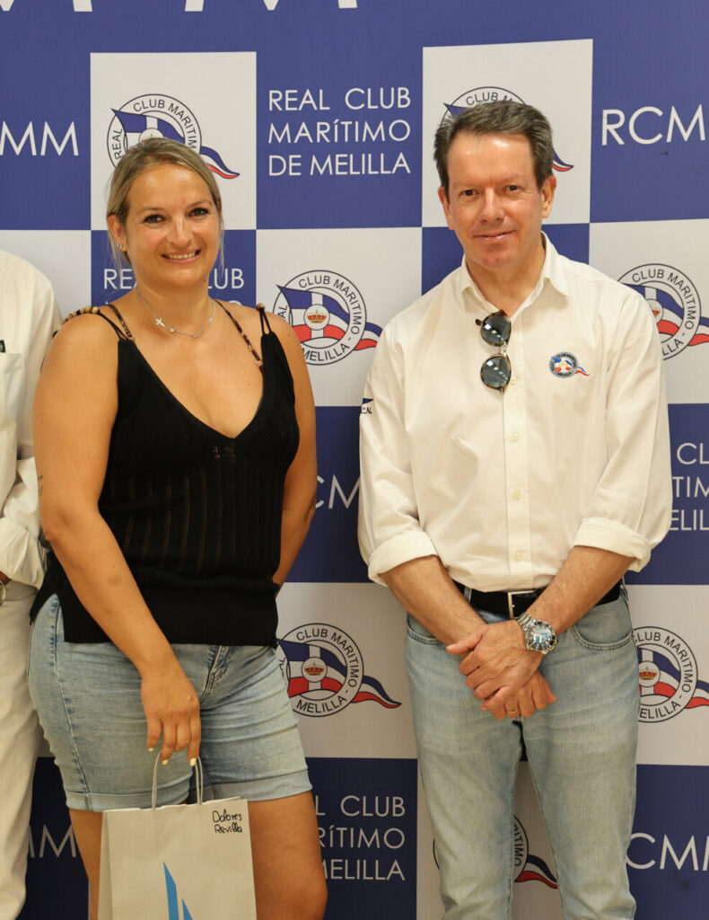 Two adults pose for a photo in front of a blue-and-white backdrop with Real Club Marítimo de Melilla logos; the woman wears a black sleeveless top and denim shorts, holding a small bag, and the man wears a white shirt with sunglasses tucked into the neckline and jeans.