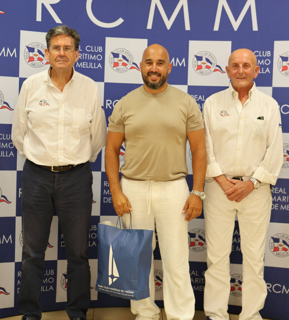 Three men pose for a photo in front of a blue-and-white branded backdrop; the center man in a beige shirt holds a blue gift bag.