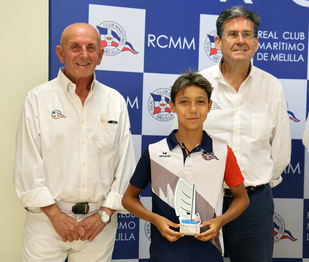 Boy in a team polo holds a sailing trophy between two men in white shirts at an awards backdrop with RCMM branding.