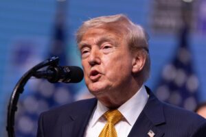 Donald Trump speaking into a microphone, wearing a dark suit and yellow tie with an American flag pin on the lapel.