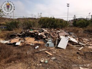 Pile of wood and drywall debris in a dry, overgrown lot with a police emblem in the upper-left corner.