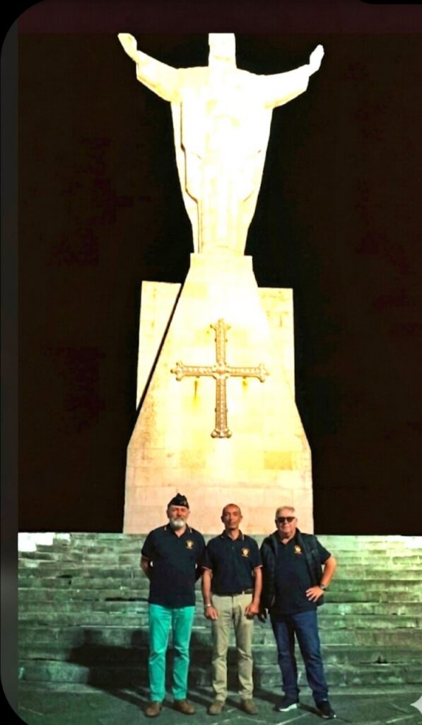 Three men stand on stone steps in front of a tall illuminated statue of Jesus with outstretched arms and a cross on the pedestal at night.