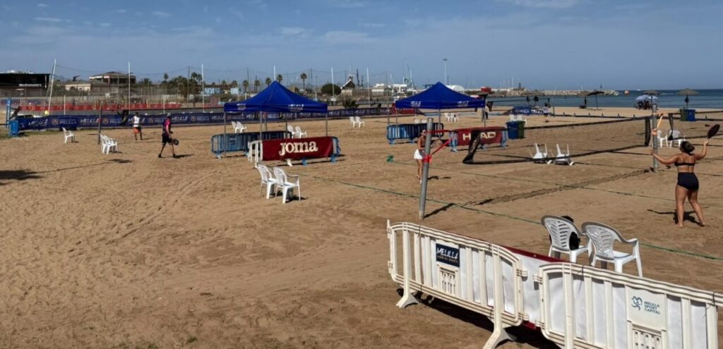 Beach volleyball court with nets and tents on sandy shore, players and chairs nearby.