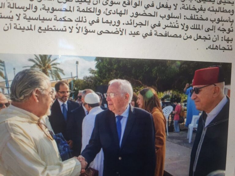 Two men shake hands at an outdoor formal event; one in a dark suit with blue tie and glasses, the other in a light shirt, with a man wearing a red fez nearby and palm trees in the background.