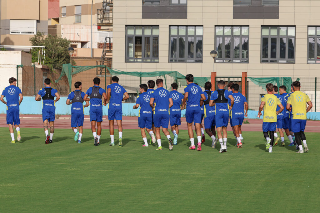 Soccer players in blue uniforms jog on a green field during practice, backs to the camera, VW logos on their shirts.