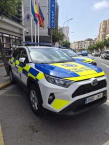 Police SUV with blue-yellow checkered livery parked on a city street in front of a Policía Local building with flags.