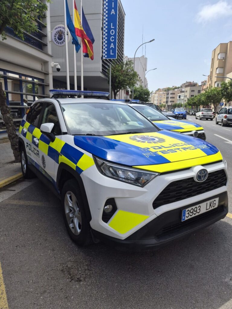 Police SUV with blue-yellow checkered livery parked on a city street in front of a Policía Local building with flags.
