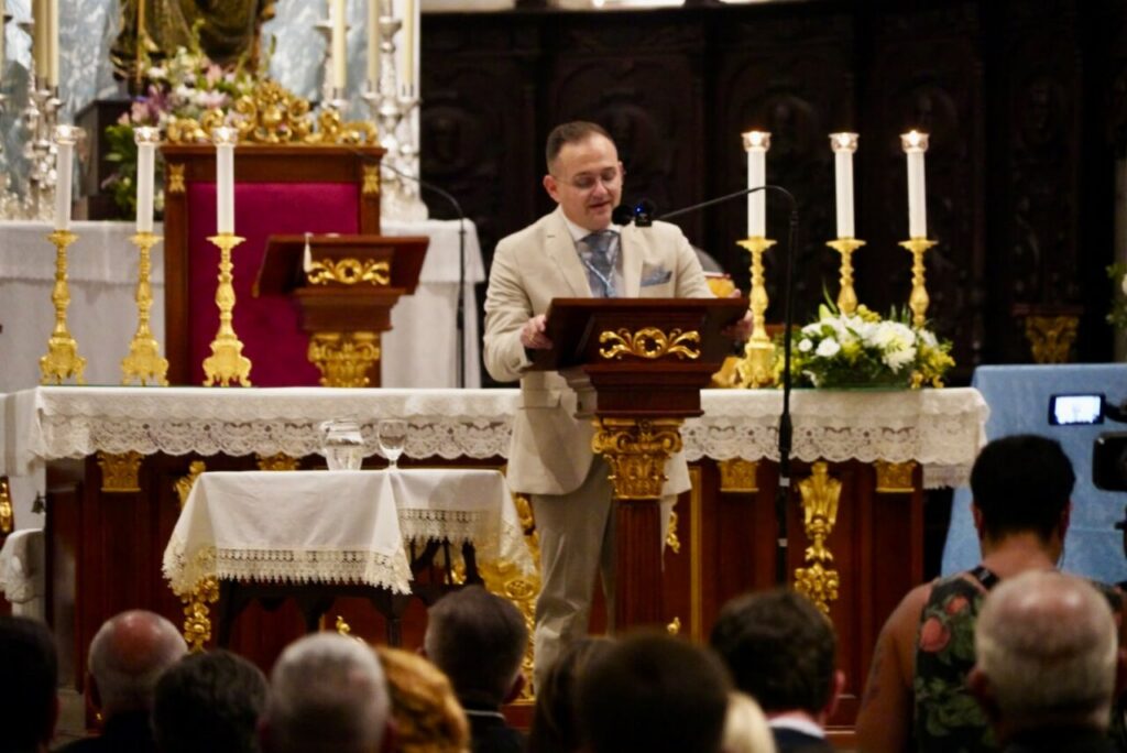 A man in a light suit speaks at a wooden lectern in a church with gold accents and white lace decorations, facing an audience.