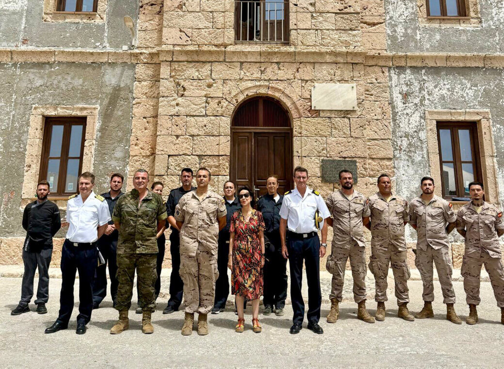 Group of military personnel in uniform standing in front of a stone building with a wooden door and arched entry. A woman in a floral dress is among them, under a bright blue sky with a lighthouse on the roof edge above.