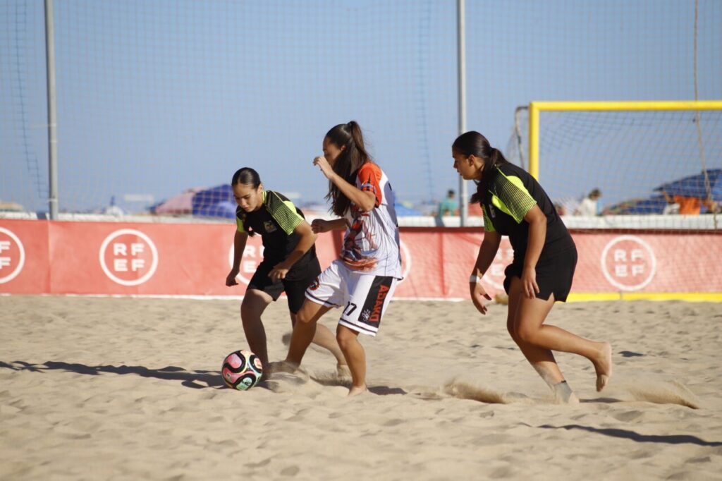 Three female beach soccer players chase a colorful ball on the sand with a goal in the background.