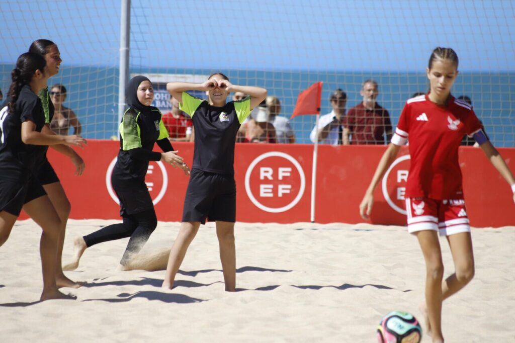 Girls playing beach soccer on a sunny day; a red-uniformed player dribbles near the ball while others in black/green chase on the sand with a red barrier and net behind.