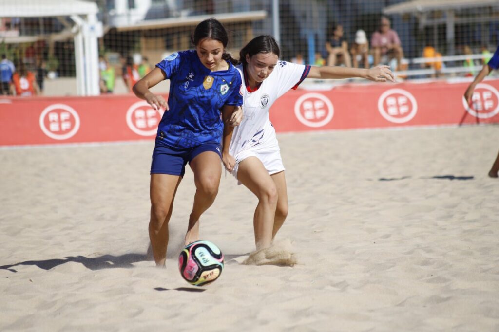 Two female soccer players in blue and white chase a colorful ball on a sandy beach during a match, with spectators in the background.