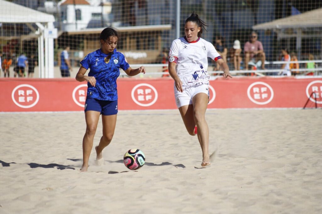 Two female soccer players on a sandy beach field chase a colorful ball during a game, with a red RF EF banner behind them.